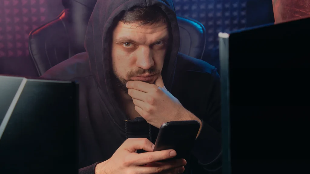 Un homme encapuchonné tient un smartphone devant ses écrans