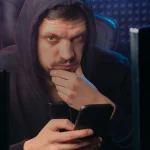 Un homme encapuchonné tient un smartphone devant ses écrans
