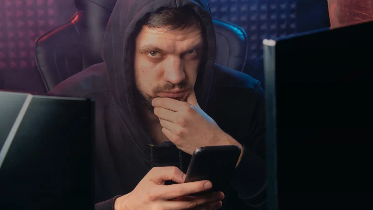 Un homme encapuchonné tient un smartphone devant ses écrans